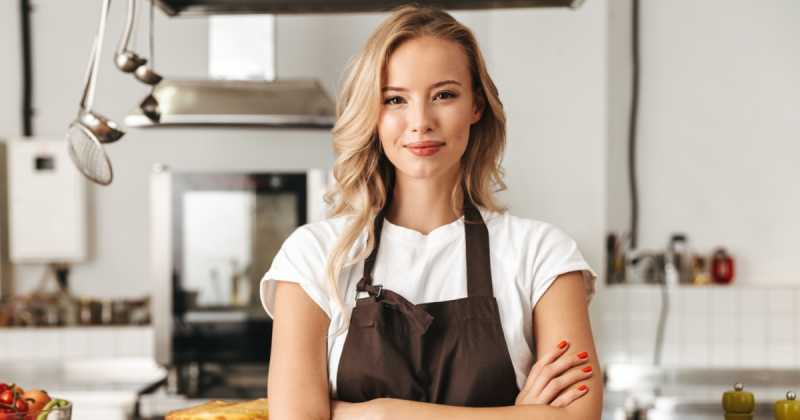 Eine selbstbewusste junge Frau in einer braunen Schürze steht mit verschränkten Armen in einer professionellen Küche.