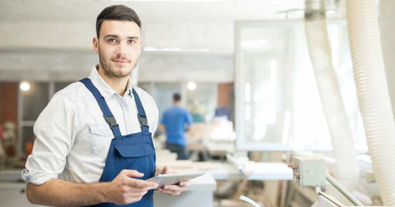 Ein Mann im Overall hält ein Tablet in der Hand und steht in einer Holzwerkstatt, im Hintergrund ist eine andere Person bei der Arbeit.