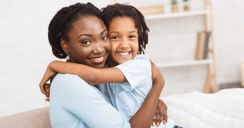 Eine fröhliche Mutter und Tochter umarmen sich und lächeln in einem Wohnzimmer in die Kamera.