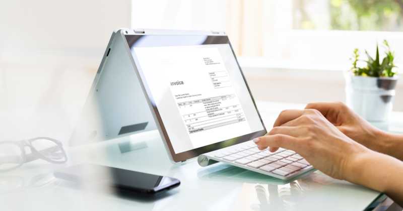 Eine Person prüft eine Rechnung auf einem Laptop-Tablet-Hybridgerät. In der Nähe stehen in einem hellen Büro ein Smartphone und eine Pflanze auf einem Glastisch.