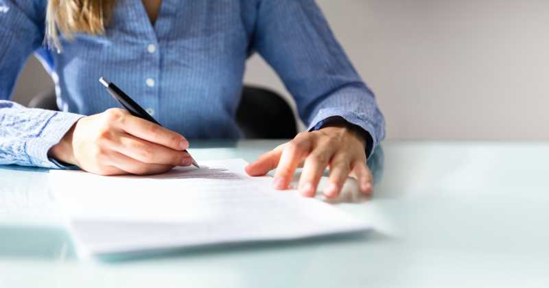 Eine Frau in einem blauen Hemd sitzt an einem Schreibtisch und schreibt mit einem schwarzen Stift auf einem Dokument.