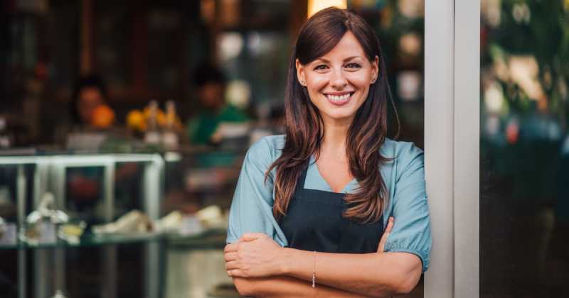 Eine lächelnde Frau mit langen Haaren, einem blauen Hemd und einer schwarzen Schürze steht mit verschränkten Armen vor einem Restaurant.