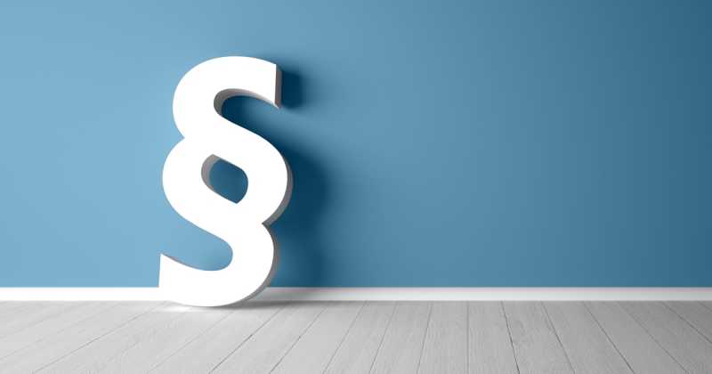 Ein großes weißes Paragraphensymbol (§), das in einem Raum mit hellem Holzboden an einer blauen Wand lehnt.