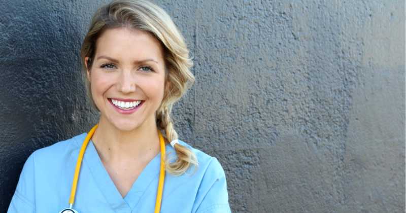 Lächelnde Frau im blauen OP-Kittel und mit Stethoskop vor einer strukturierten grauen Wand.