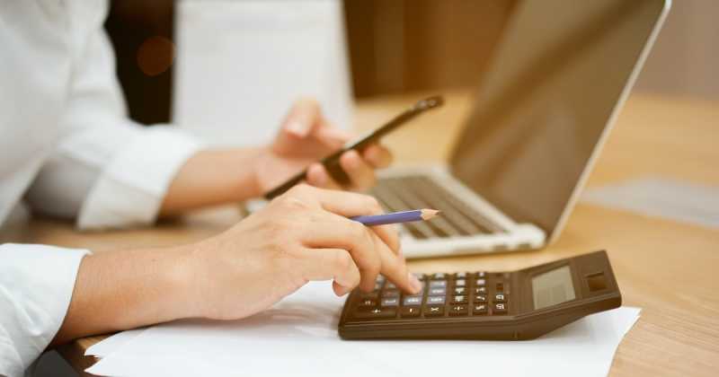 Eine Person berechnet in einem Büro mithilfe eines Taschenrechners und eines Laptops die Ausgaben.