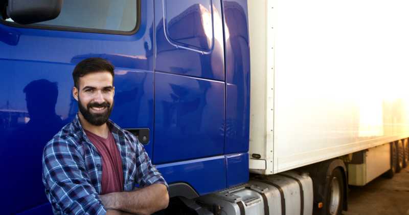 Ein lächelnder LKW-Fahrer lehnt an einem blauen LKW, im Hintergrund geht die Sonne unter.
