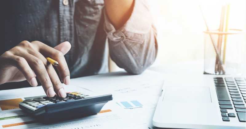 Eine Person, die einen Taschenrechner neben einem Laptop und Finanzdokumenten auf einem Schreibtisch verwendet, was darauf hinweist, dass sie eine Finanzplanung oder ein Budget durchführt.