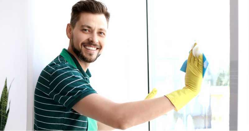Ein Mann in einem gestreiften Hemd und gelben Handschuhen lächelt, während er in einem hellen, sonnendurchfluteten Raum ein Fenster putzt.