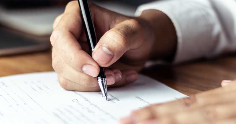 Eine Person im weißen Hemd unterschreibt ein Dokument mit einem schwarzen Stift und konzentriert sich dabei voll und ganz auf ihre Aufgabe.