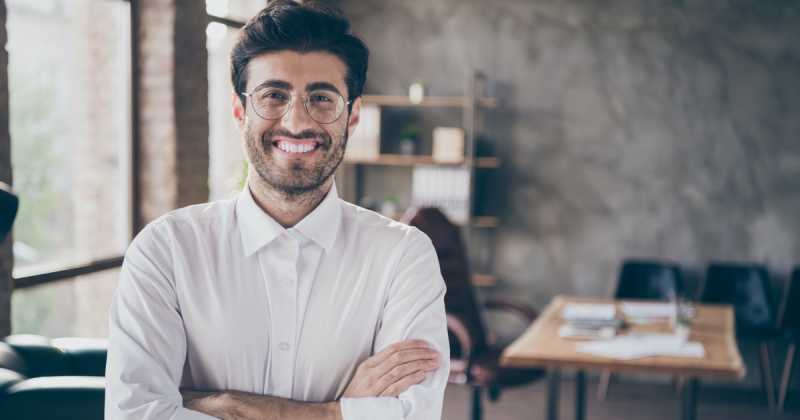 Lächelnder Mann mit Brille, weißem Hemd und verschränkten Armen steht in einer Büroumgebung.