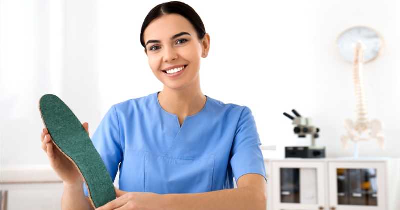 Eine Podologin im blauen Kittel lächelt und hält eine orthopädische Einlegesohle in einer Klinik mit medizinischen Geräten im Hintergrund.
