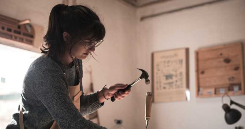 Eine Frau untersucht in einer Werkstatt einen kleinen Hammer, im Hintergrund sind Holzbearbeitungswerkzeuge und ein Poster zu sehen.