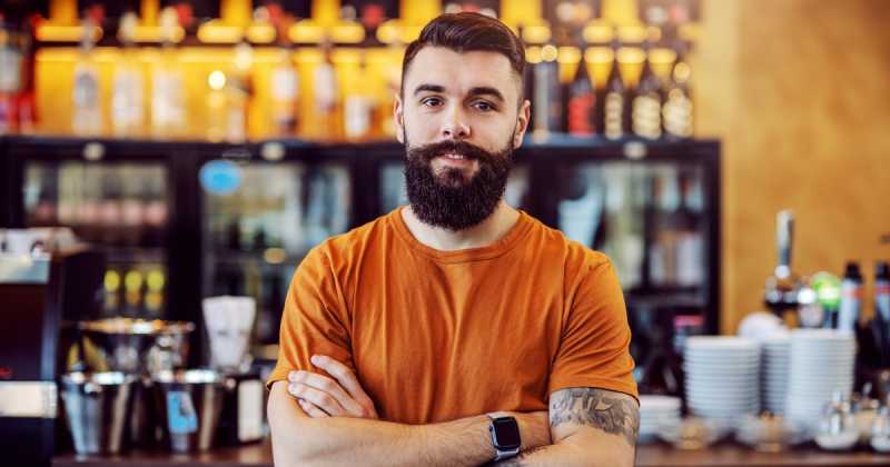 Mann mit Bart und Tattoos steht mit verschränkten Armen vor einer Bar und trägt ein orangefarbenes T-Shirt.