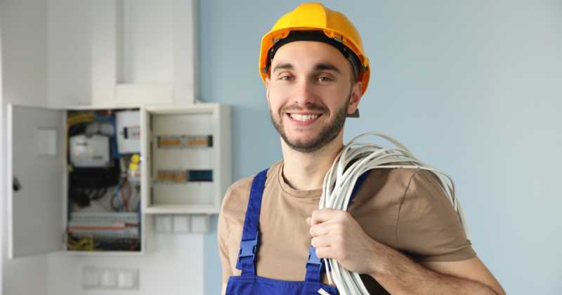 Lächelnder Elektriker mit gelbem Schutzhelm und Kabeln in der Hand, der vor einem offenen Schaltschrank steht.