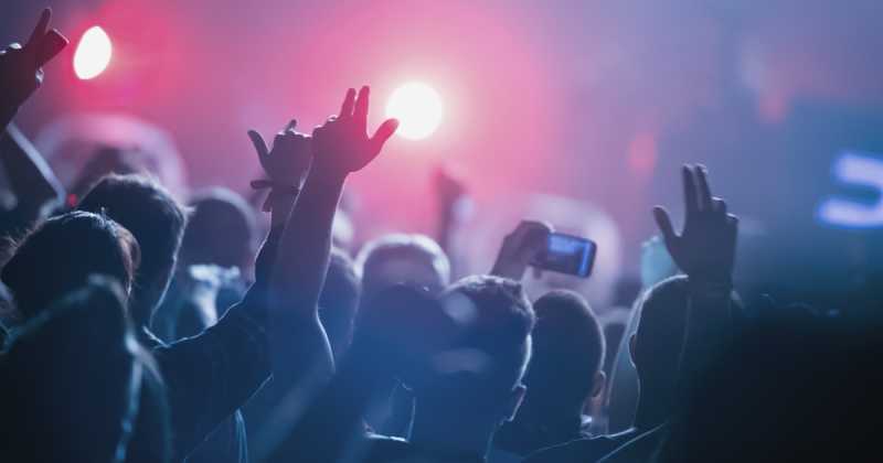 Eine Menschenmenge bei einem Konzert mit erhobenen Händen, die sich vor hellem Bühnenlicht und blauem Dunst abzeichnen.