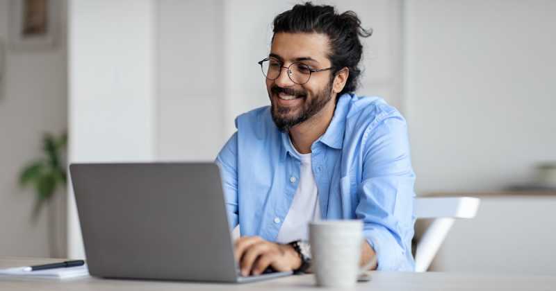 Ein lächelnder Mann mit Brille und Bart, der ein blaues Hemd trägt und an einem weißen Schreibtisch an einem Laptop arbeitet.
