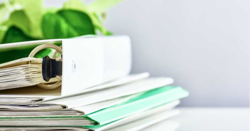 Stapel weißer und grüner Ordner mit Dokumenten auf einem Schreibtisch, Fokus auf den Metallringen des offenen Ordners.