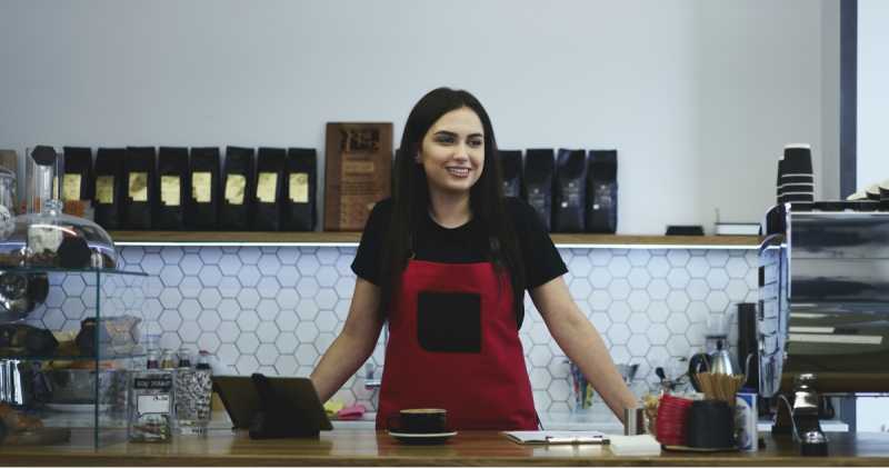 Ein lächelnder Barista in einer roten Schürze steht in einem modernen Café hinter der Theke, umgeben von verschiedenen Kaffeezubereitungsgeräten.