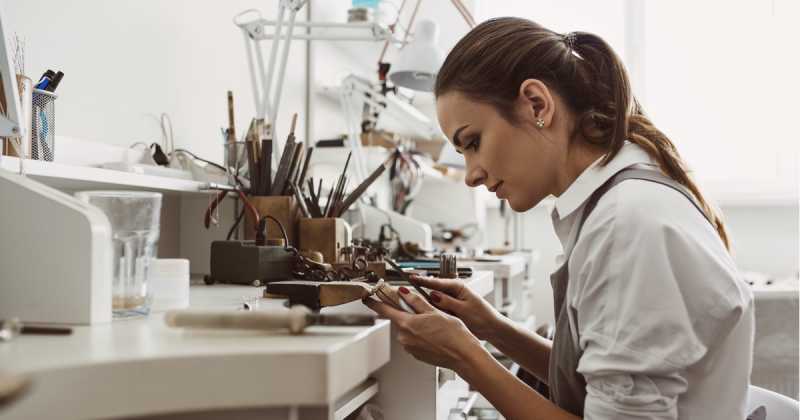 Eine Frau im weißen Hemd arbeitet konzentriert an einer Schmuckwerkbank, verwendet Werkzeuge und untersucht ein Stück genau.