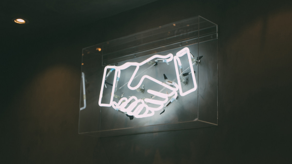 Ein Zeichen aus Neonröhren. Es zeigt einen Handschlag oder Händedruck.