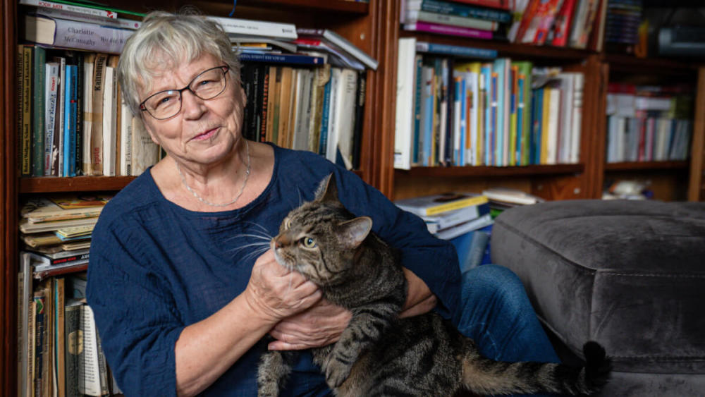 Eine ältere Frau sitzt auf dem Boden und lehnt sich an ihr Bücherregal. Auf ihrem Schoß sitzt ihre Katze. 