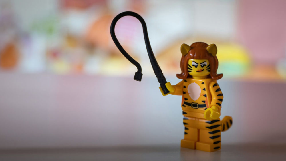 Eine weiblich gelesene Lego-Figur hält eine Peitsche und blickt herausfordernd.