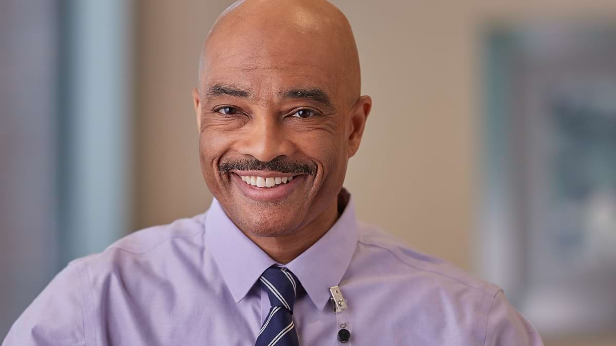 A person with a moustache smiling at the camera. They are wearing a lilac shirt and a name badge. 