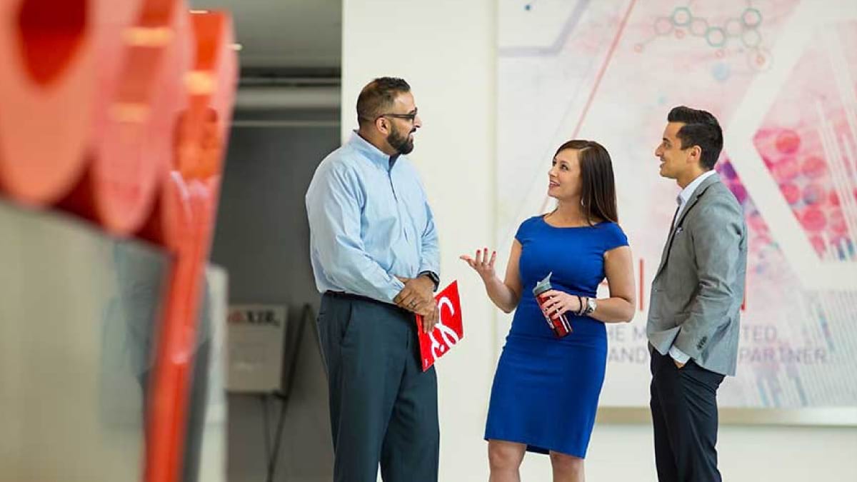 Three professionals standing up having a conversation indoors at a Lilly facility. One person holding a Lilly brochure, another holding a water bottle. 