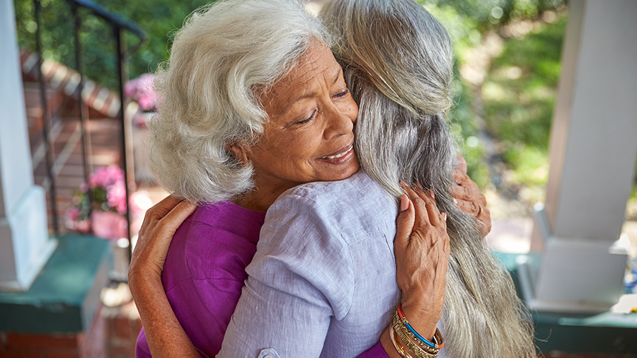 Two people joyfully embracing while standing on a porch. Both have grey hair, one person is wearing a purple top and the other person is wearing a grey shirt. 