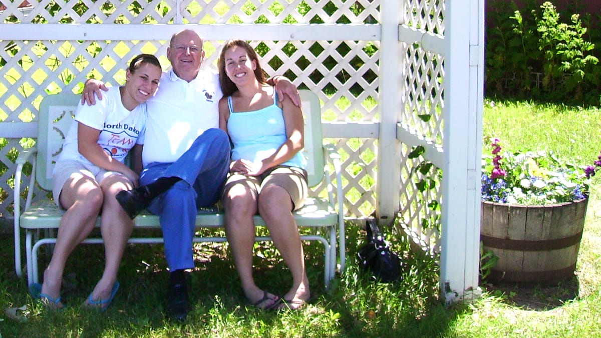 Three adults sitting on an outdoor bench in the sun. They are smiling and embracing. 