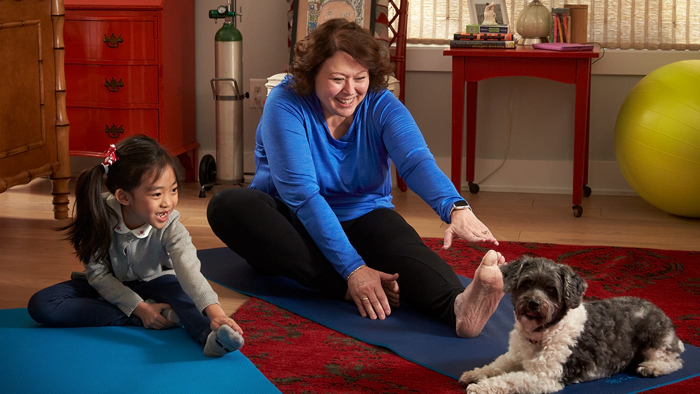 An adult and a young child smiling, sitting down and stretching on yoga mats with an exercise ball in the background. A small dog is nearby.