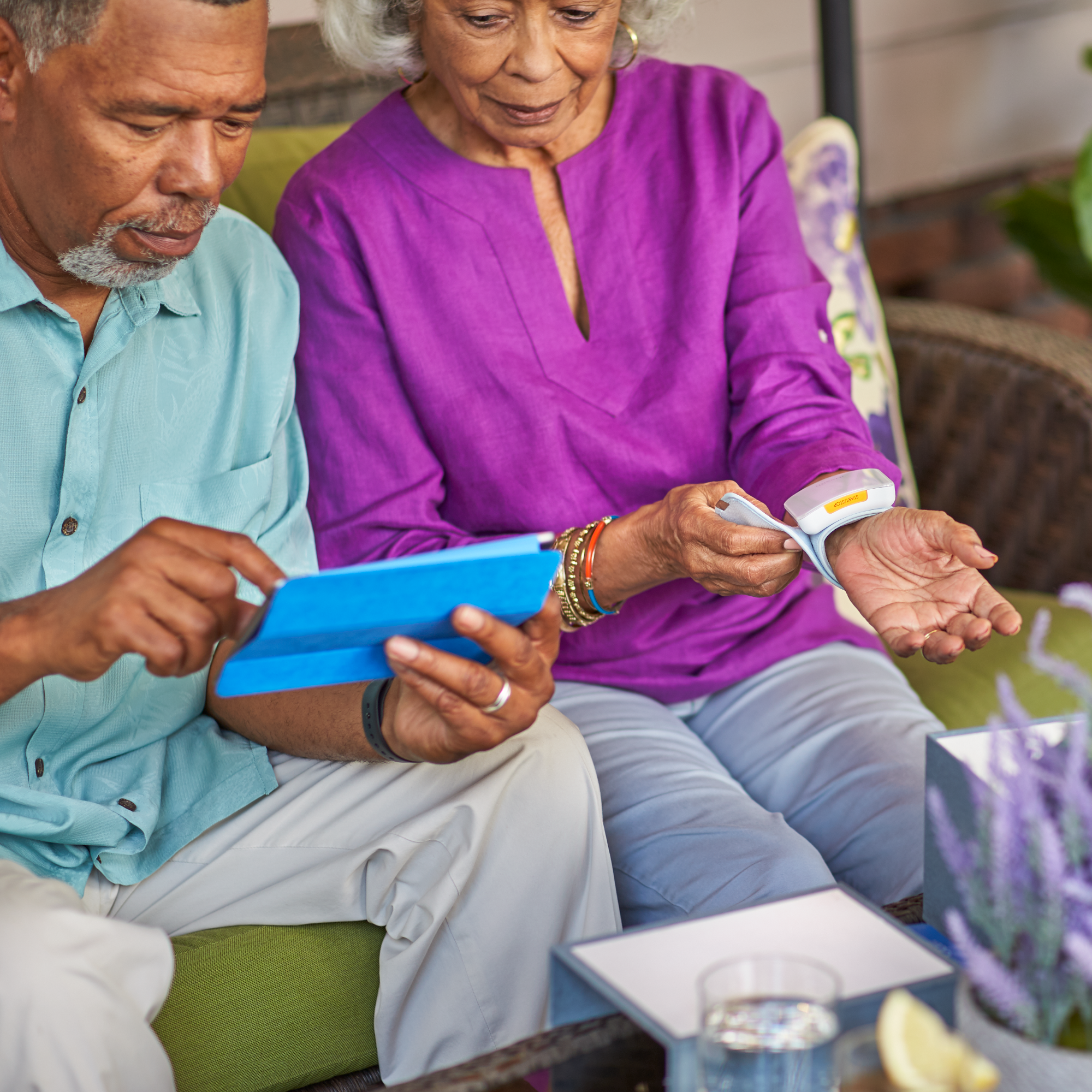 Two adults sitting side by side. One person is applying a medical device to their wrist, the other is looking at a handheld tablet device.
