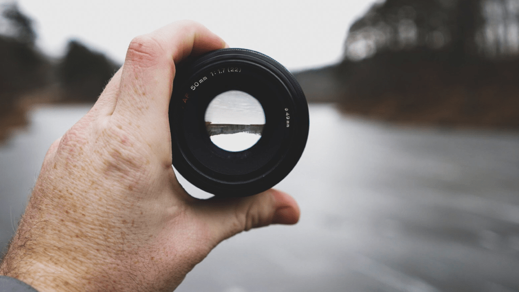 A camera lens held in a persons left hand. It is focused on an outdoor landscape in the distance, indicating the future.  