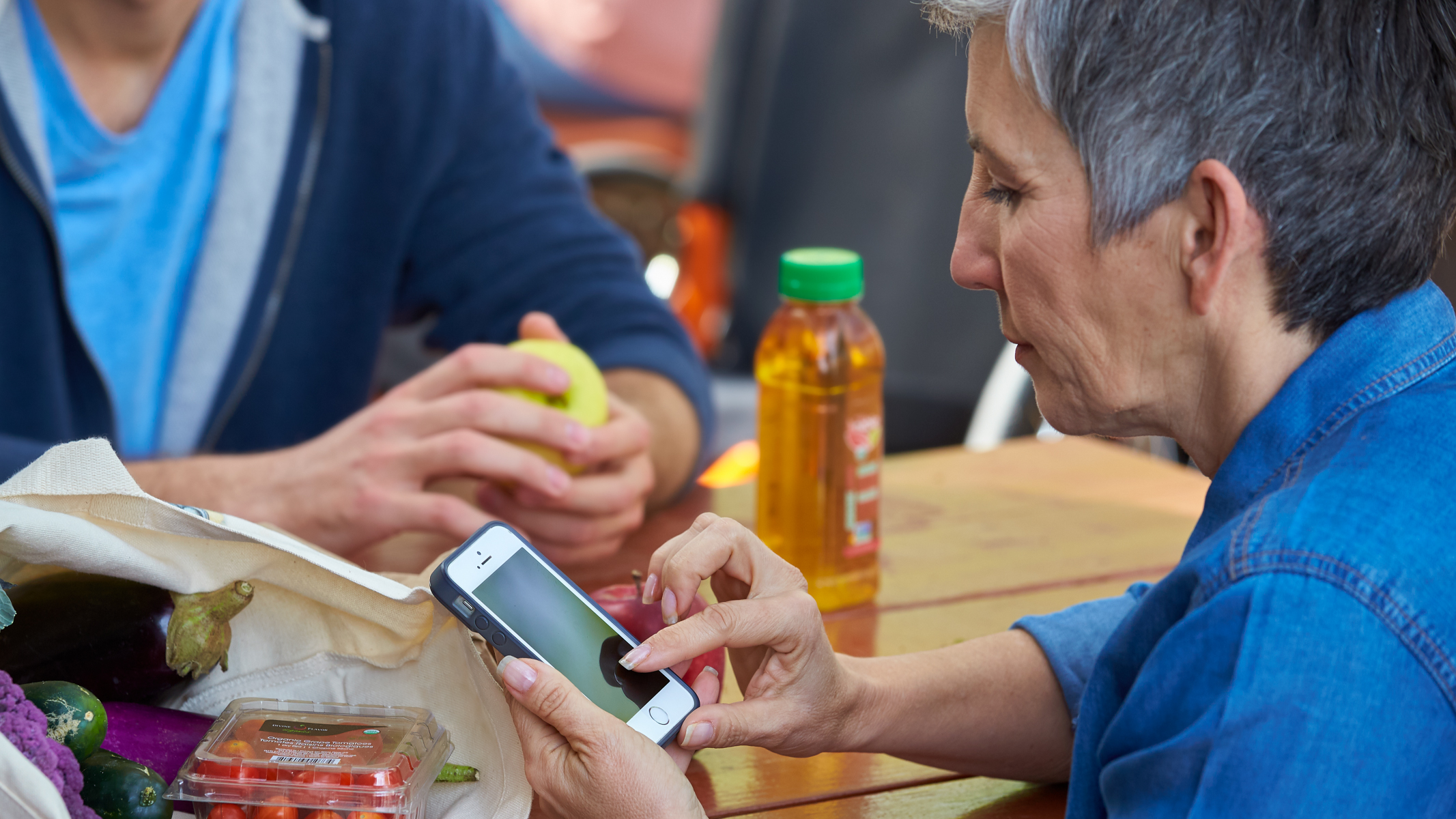 An adult scrolling on their phone while sitting at a picnic table with another person. A bag of fresh vegetables is on the table.