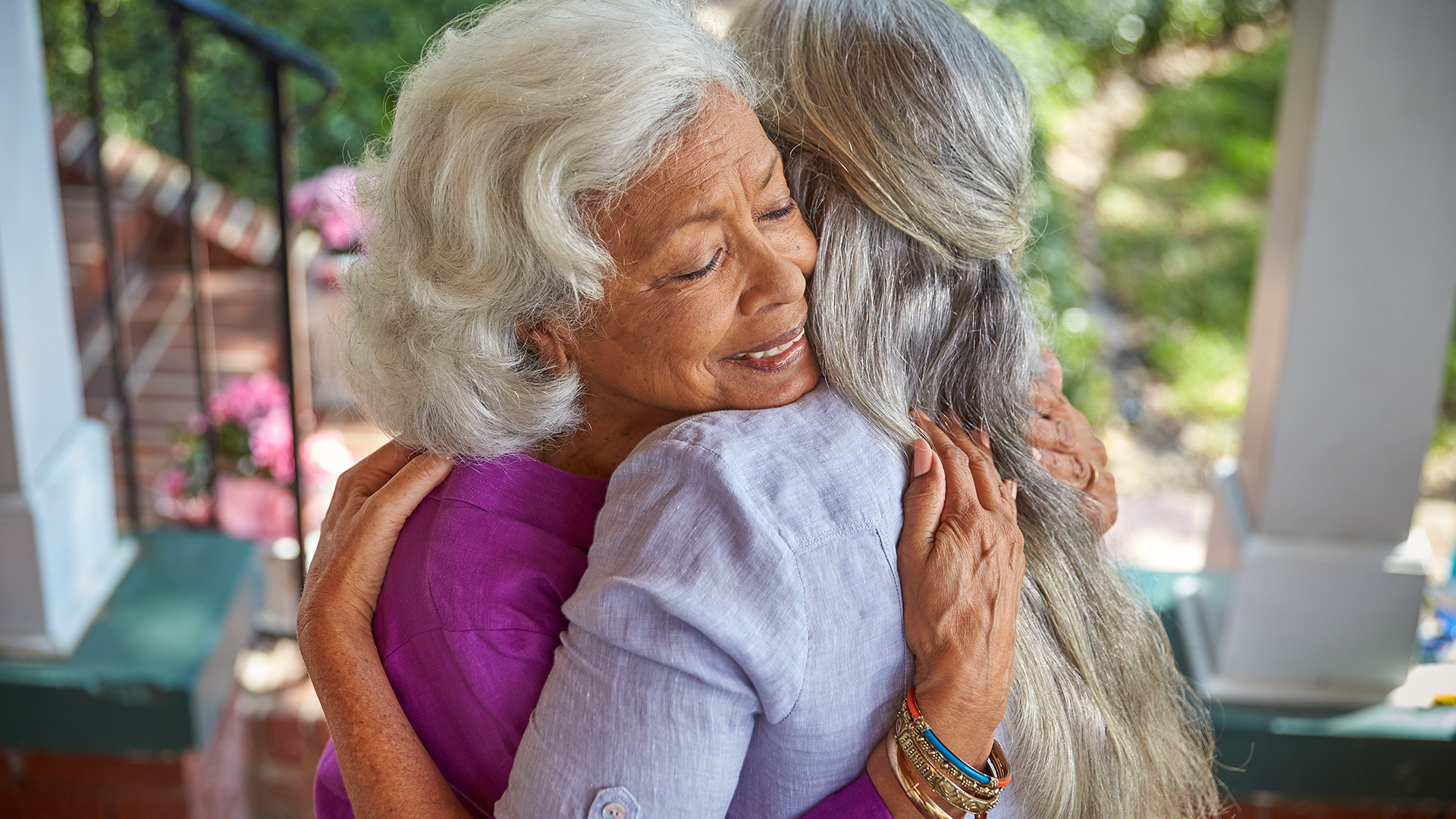 Two people joyfully embracing while standing on a porch. Both have grey hair, one person is wearing a purple top and the other person is wearing a grey shirt.