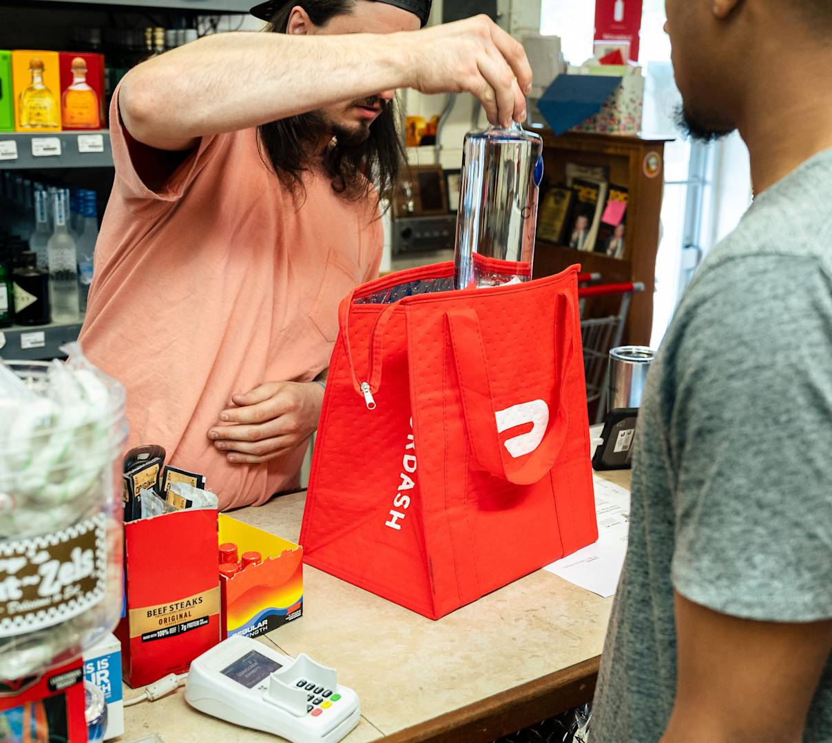 Employee at Corkscrew Johnny's place liquor bottle in DoorDash bag for Dasher at register