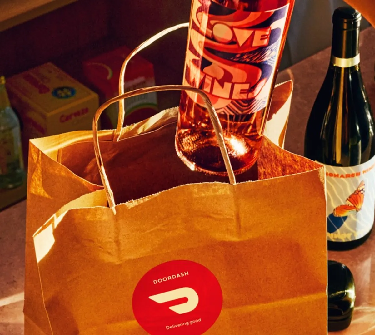 Brown DoorDash bag with wine bottles inside, surrounded by additional wine bottles on a counter in warm lighting.