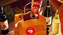 Brown DoorDash bag with wine bottles inside, surrounded by additional wine bottles on a counter in warm lighting.