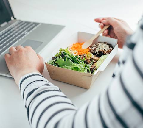 Person in striped shirt eating delivered lunch bowl with salad, rice, and protein while working on laptop.