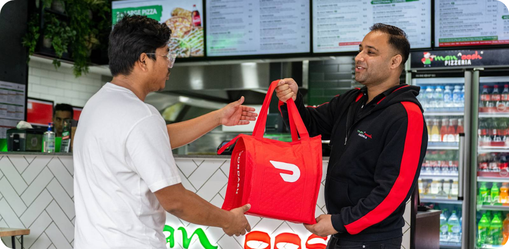 Manoosh Pizzeria employee hands a Dasher a DoorDash bag