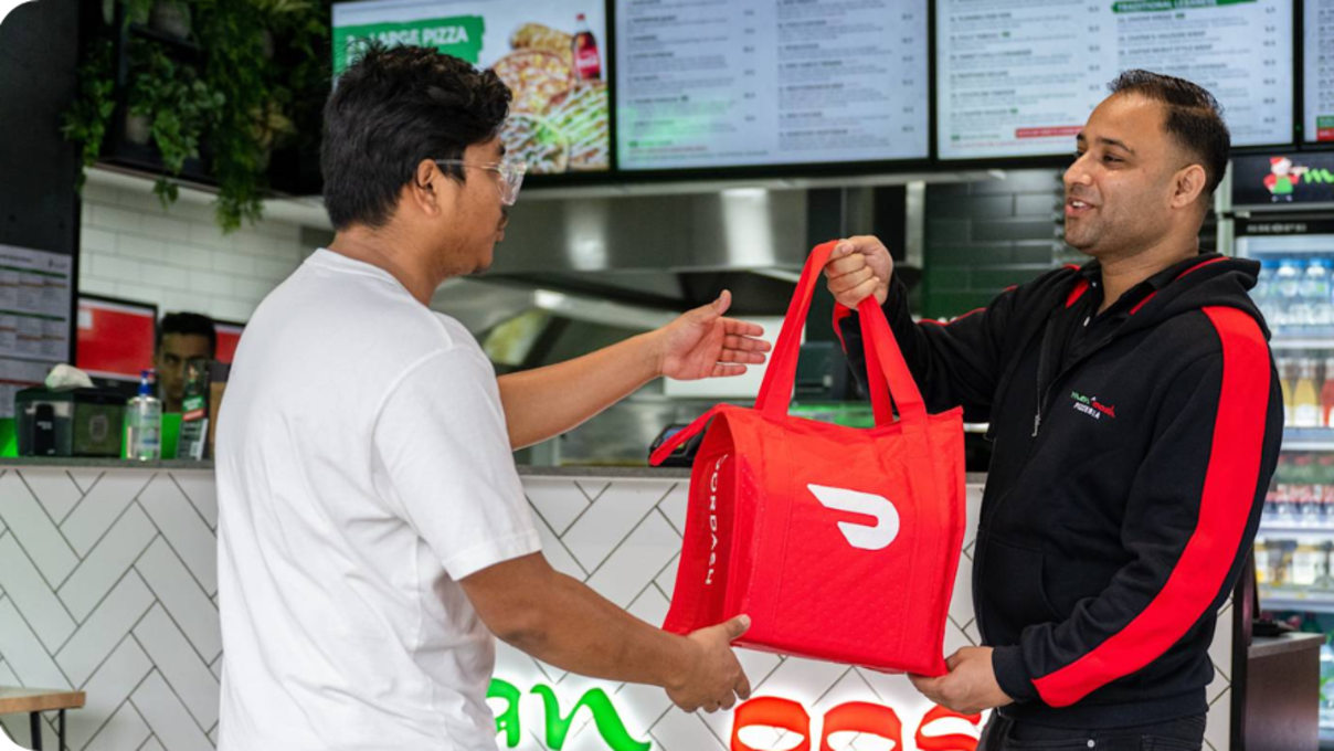 Manoosh Pizzeria employee hands a Dasher a DoorDash bag