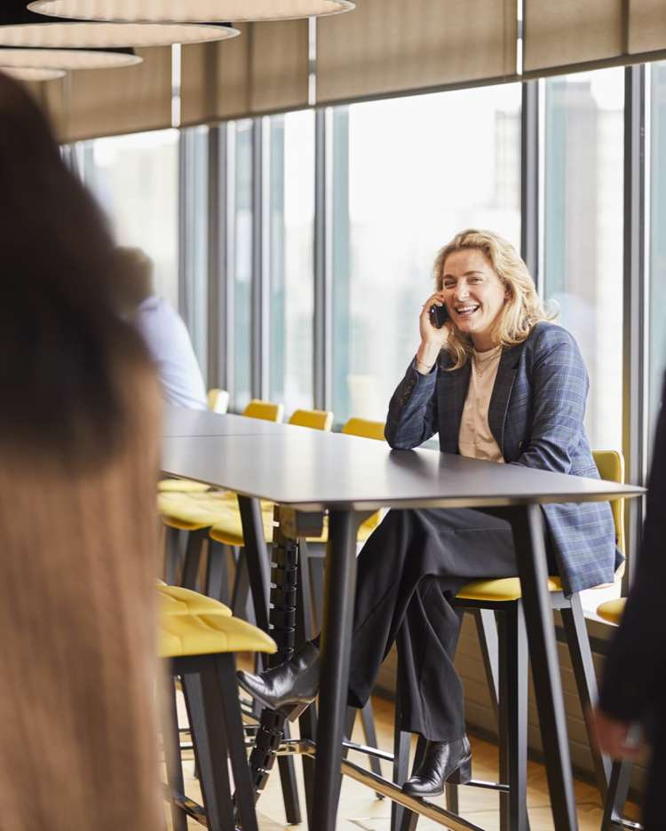 professional woman talking on the phone at a tall table with windows behind her looking out on an urban scene