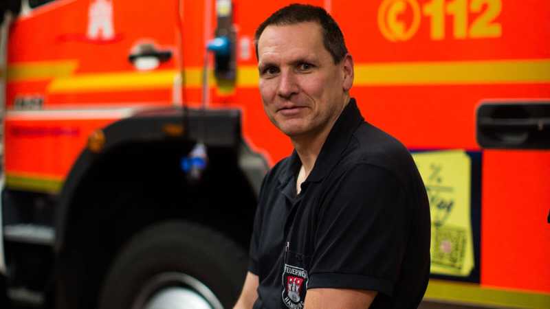 Das Bild zeigt einen Feuerwehrmann der vor einem Feuerwehrfahrzeug sitzend seitlich in die Kamera schaut.