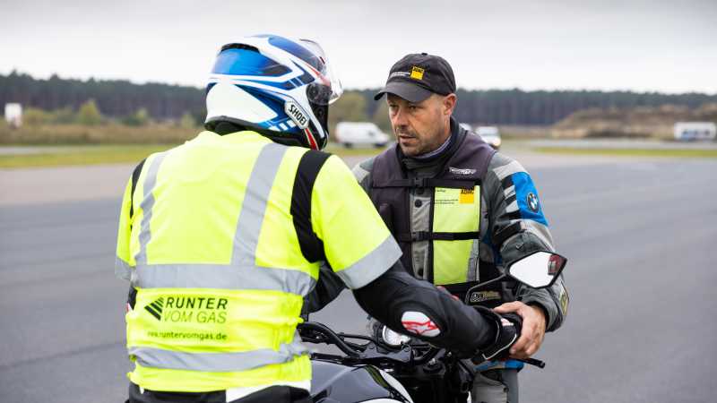 Der Trainer steht vor Teilnehmer Dirk und seinem Motorrad und zeigt ihm die Handhaltung zum Bremsen.