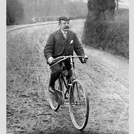 Historische schwarz-weiß Aufnahme eines Mannes mit Schnurrbart, der ein Fahrrad fährt.