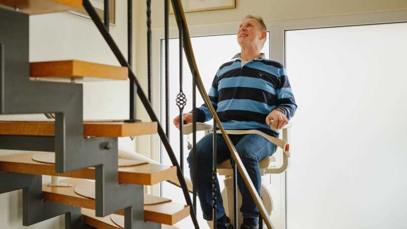 Ein Mann sitzt auf dem Stuhl eines Treppenliftes und blickt nach oben. 