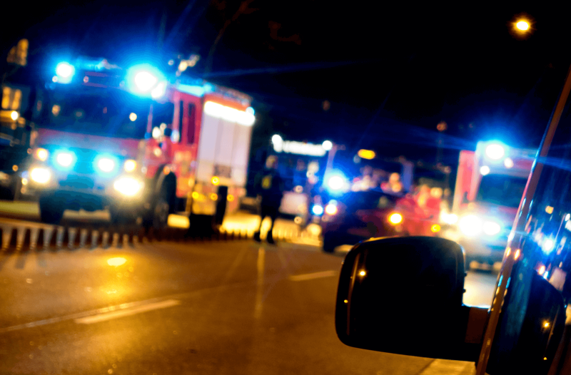 Man sieht eine Unfallstelle bei Nacht: Ein Feuerwehrauto und Krankenwagen stehen neben einem Auto mit Blaulicht. Die Licht verschwimmen.