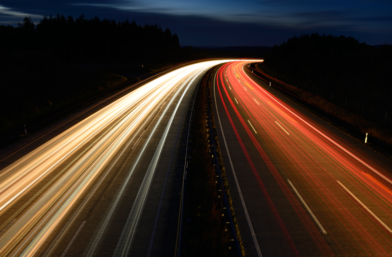 Das Bild zeigt eine inszenierte Fotografie mit ineinander übergehenden Lichteffekten von schnell fahrenden Autos auf einer Autobahn bei Nacht.