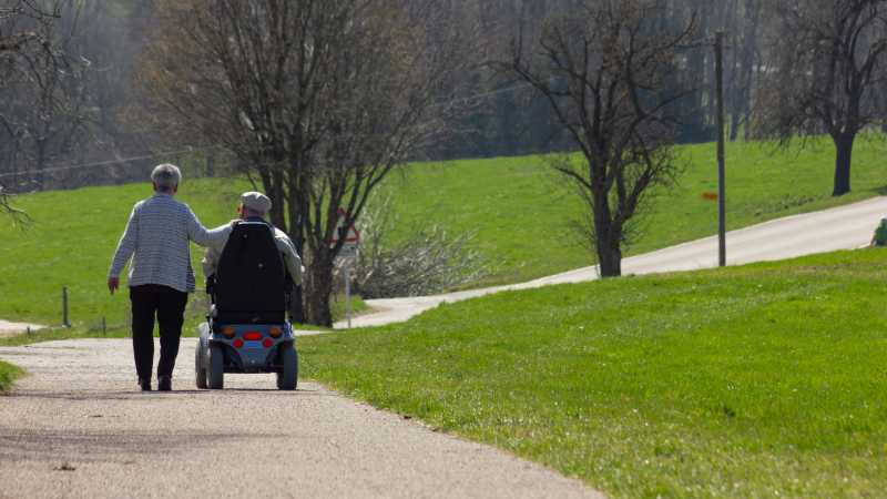 Ein älteres Paar geht in einem Park spazieren. Der Mann sitzt dabei in einem motorisierten Krankenfahrstuhl.