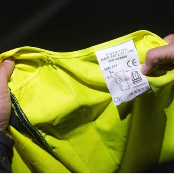 Eine Hand zeigt ein weißes Etikett einer neongelben Warnweste aus
fluoreszierendem Material.